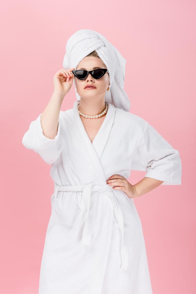 junge übergroße Frau im Bademantel, Sonnenbrille und Handtuch auf dem Kopf stehend mit der Hand auf der Taille isoliert auf rosa  - Foto, Bild