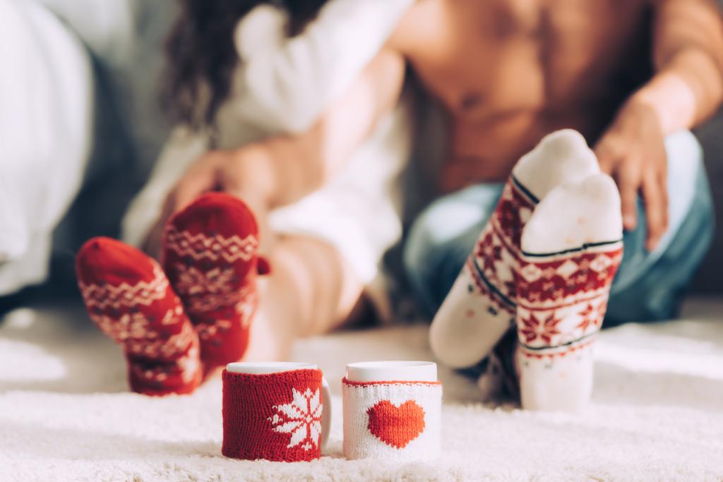 selective focus of cups of coffee with woolen cases and legs of couple sitting on floor at home - Photo, Image