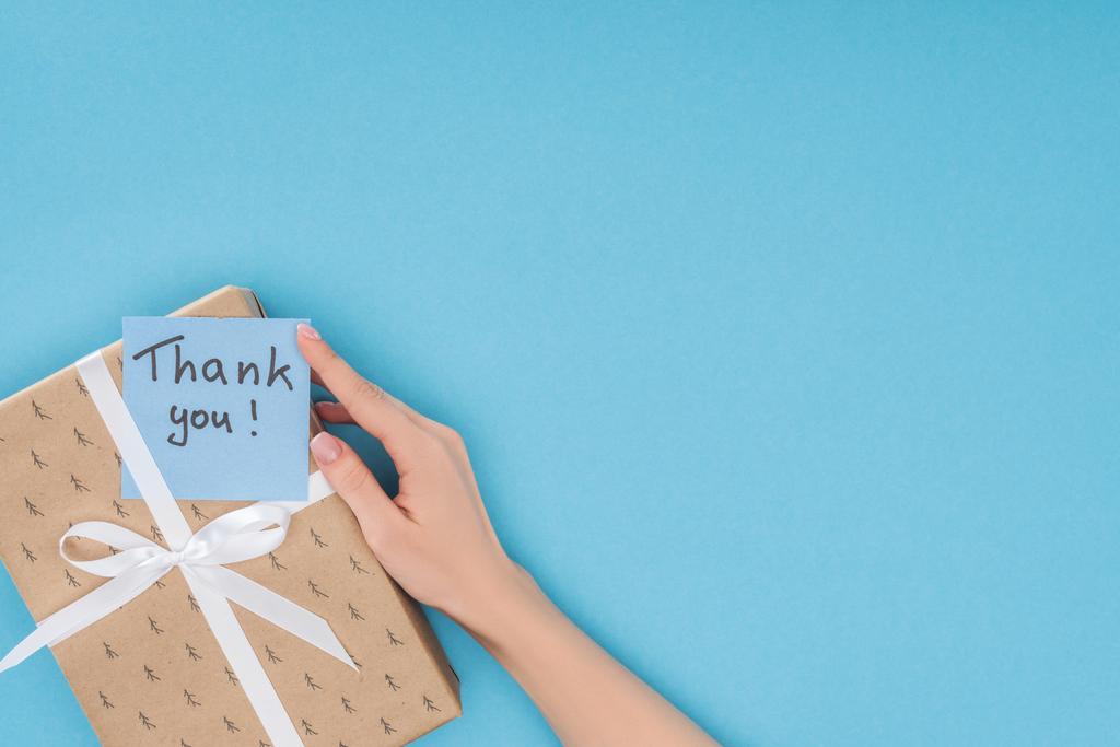 vista recortada de la mano y la nota adhesiva con letras de agradecimiento en la caja de regalo aislado sobre fondo azul
  - Foto, Imagen