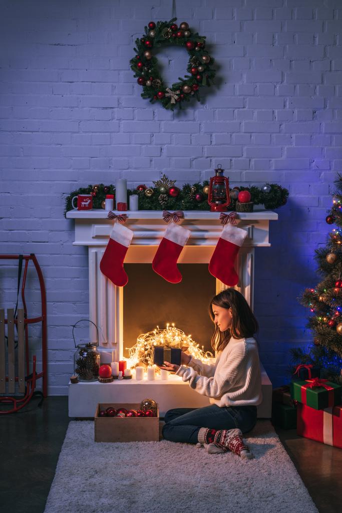 schöne Frau hält Geschenk und sitzt in der Nähe von Weihnachten dekorierten Kamin und Weihnachtsbaum - Foto, Bild
