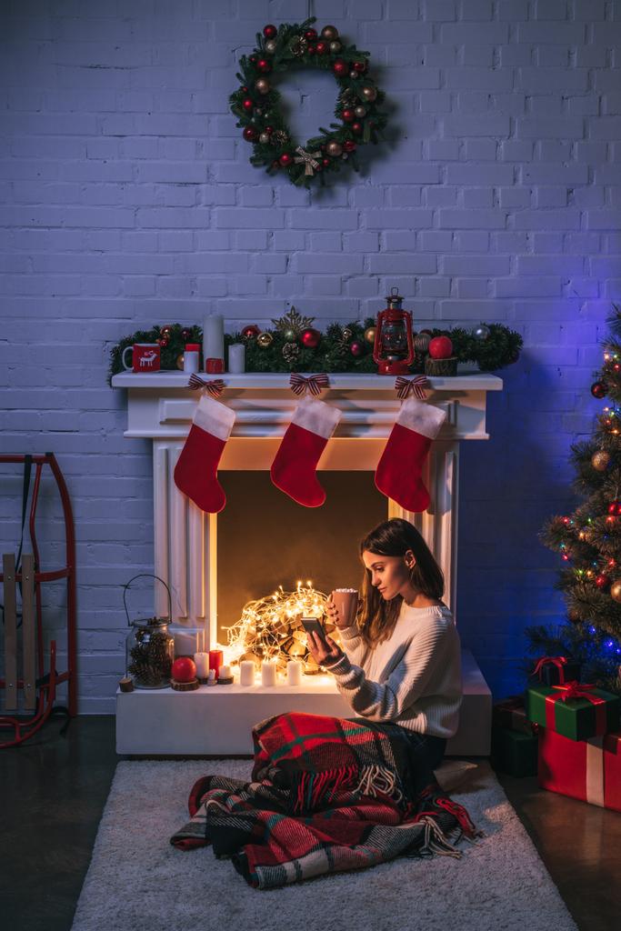 ελκυστική γυναίκα χρησιμοποιώντας το smartphone κοντά Χριστούγεννα διακόσμηση, τζάκι και το χριστουγεννιάτικο δέντρο - Φωτογραφία, εικόνα