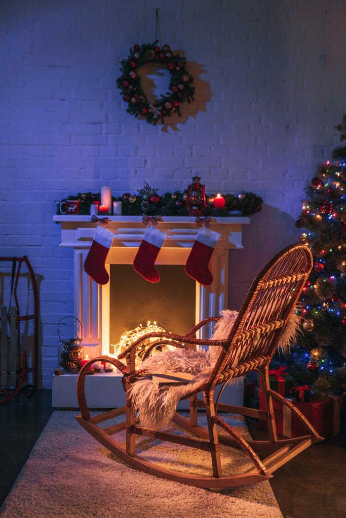 Open haard met kerstversiering in de buurt van de kerstboom en houten schommelstoel  - Foto, afbeelding