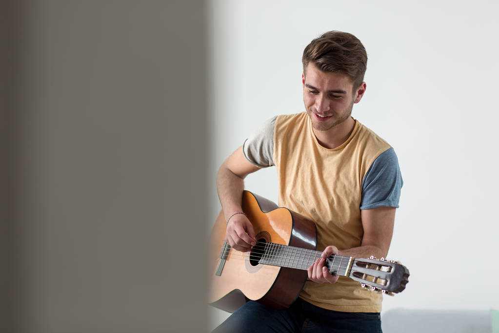 Pretty young man playing guitar while sitting on sofa in light living room - Photo, Image