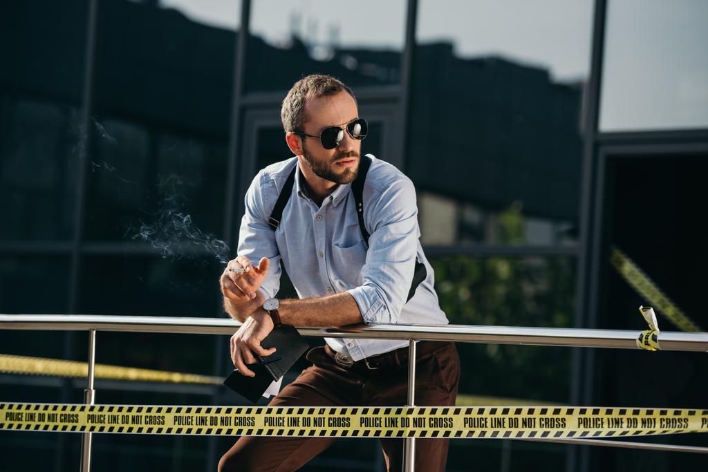 чоловічий детектив в сонцезахисних окулярах курить і дивиться на бік, спираючись на паркан
 - Фото, зображення