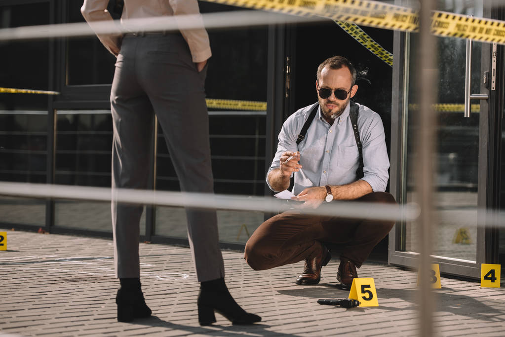 sitting male detective in sunglasses smoking and looking at crime scene with standing partner near him - Photo, Image