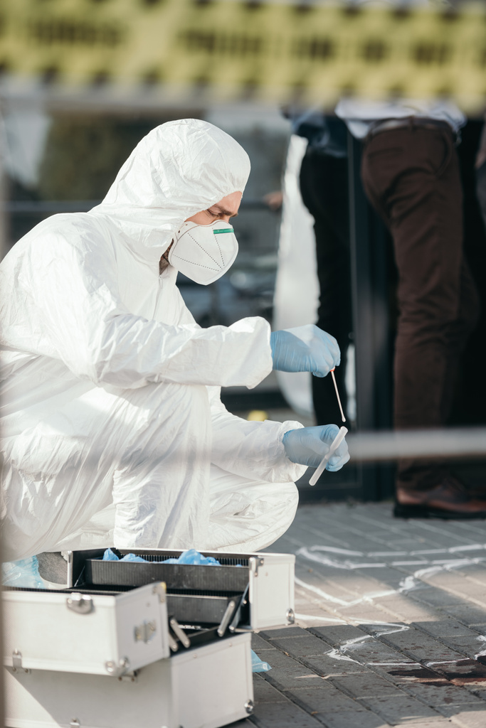 Kriminologe in Schutzanzug und Latexhandschuhen entnimmt Blutprobe im Reagenzglas am Tatort - Foto, Bild