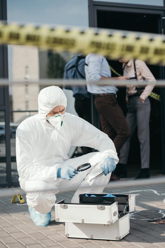 mannelijke criminoloog in beschermend pak en latex handschoenen kijken door een vergrootglas in belangrijk bewijs - Foto, afbeelding