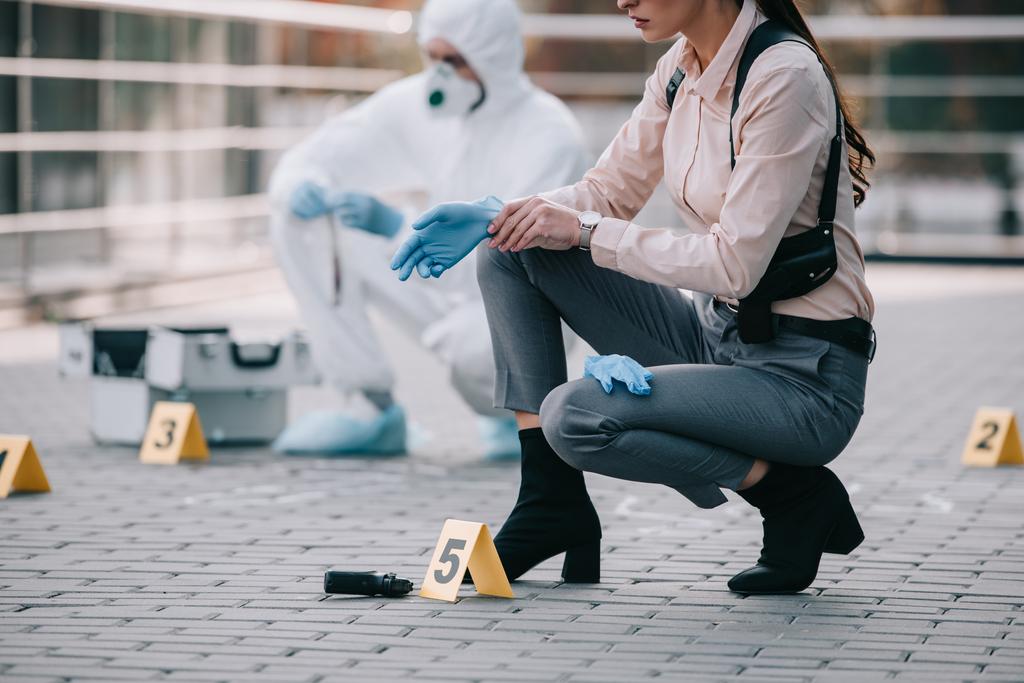 cropped view of female detective wearing latex gloves with criminologist behind at crime scene - Photo, Image