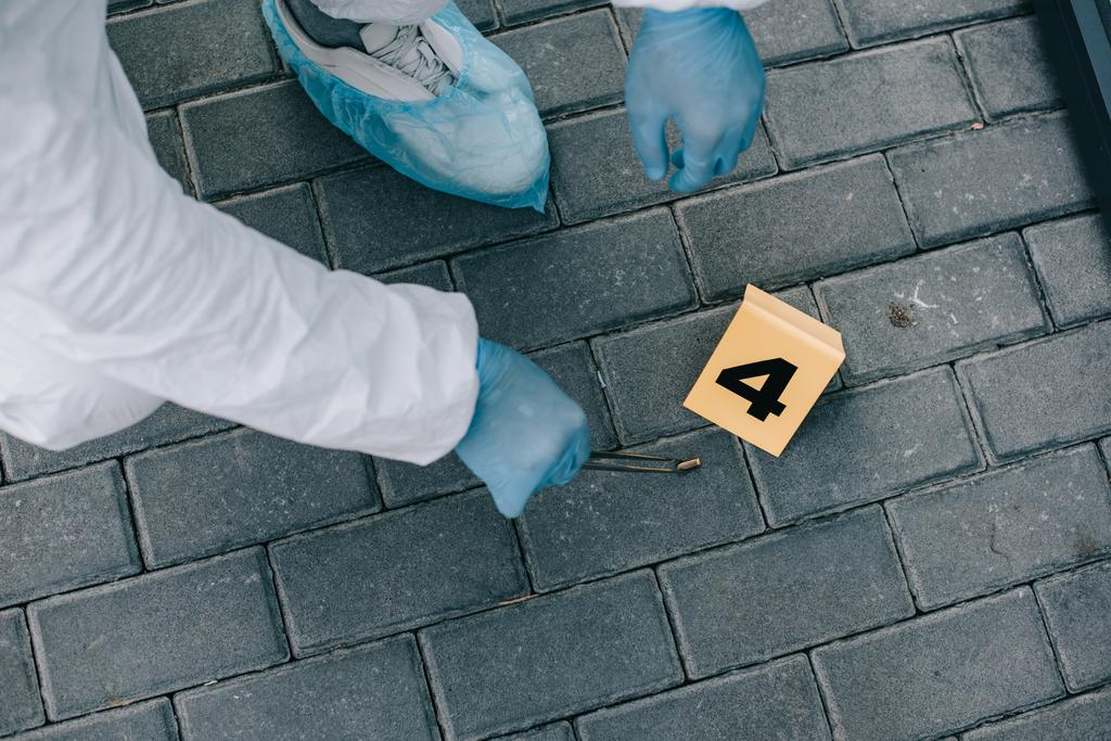 Teilansicht eines Kriminologen in Schutzanzug und Latexhandschuhen, der am Tatort mit einer Pinzette Beweismittel in Fläschchen steckt - Foto, Bild