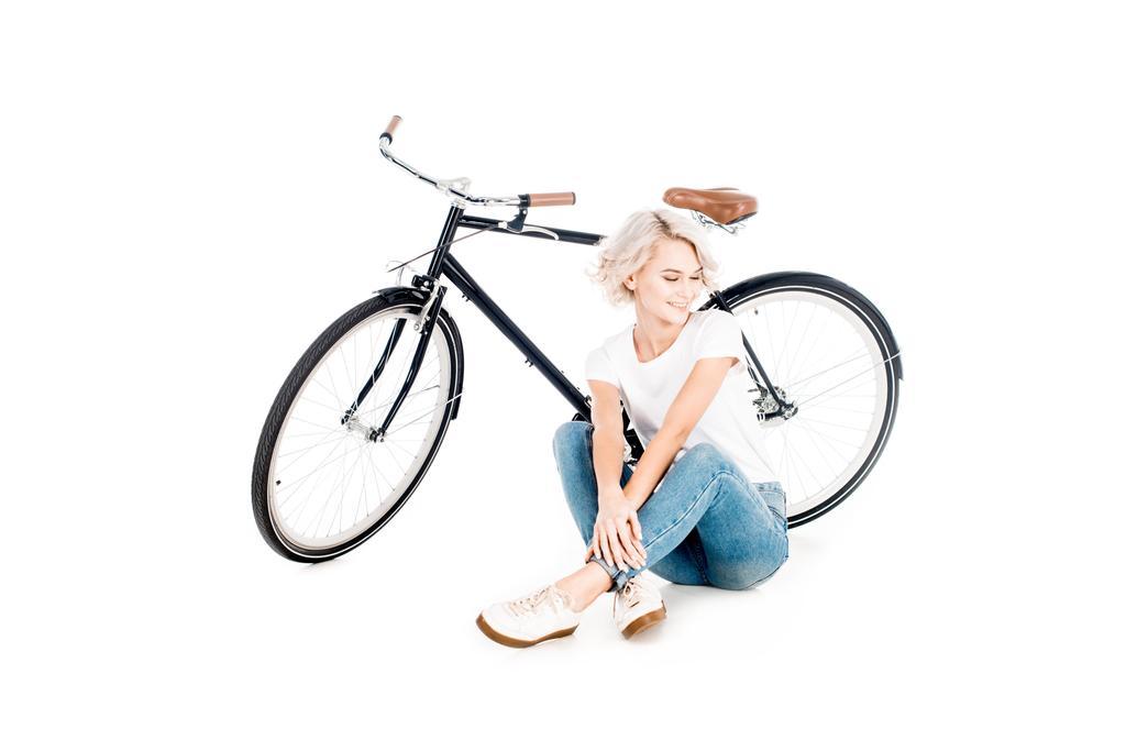ホワイトに分離された自転車のそばに座って素晴らしい金髪の若い成人女性 - 写真・画像