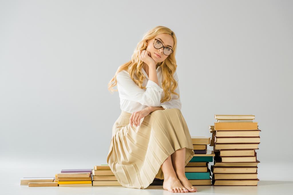 attractive thoughtful barefoot woman in glasses sitting on steps made of books   - Photo, Image