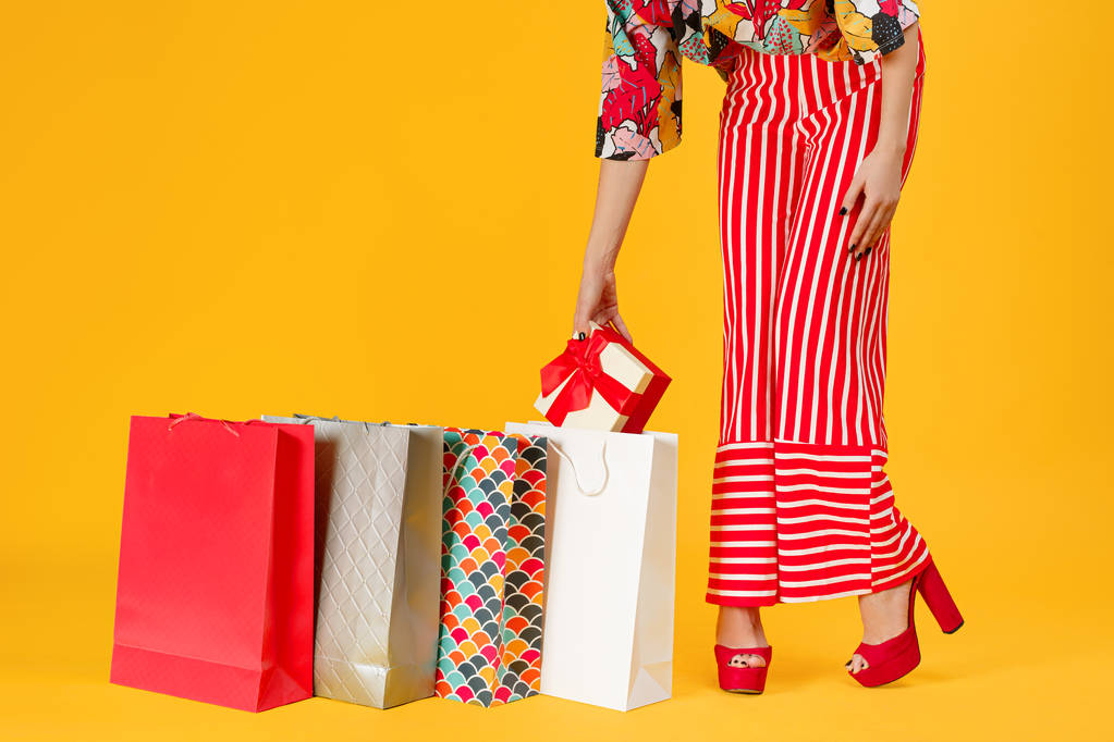 Imagen recortada de una mujer en pantalones rojos y sandalias sosteniendo una caja de regalo con coloridas bolsas de compras sobre un fondo naranja
. - Foto, imagen