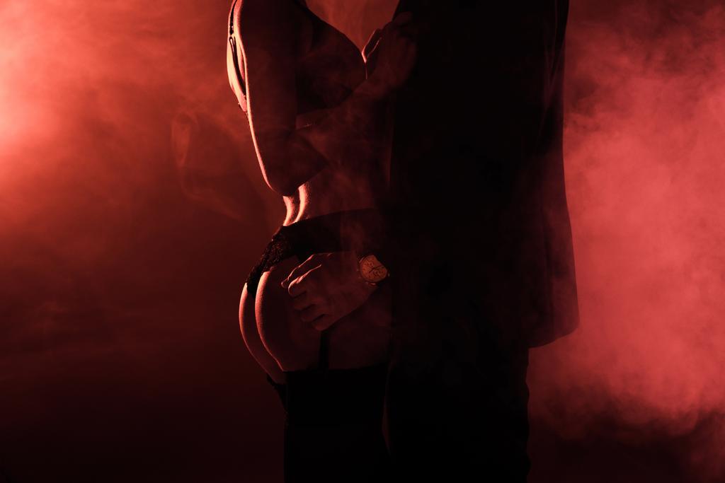 Vista parziale di attraente coppia seducente in abbraccio su sfondo fumo rosso
 - Foto, immagini