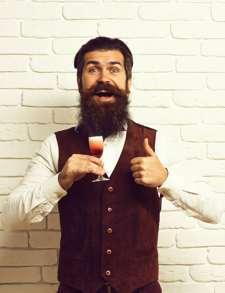красивый бородатый мужчина с длинной бородой и усами имеет стильные волосы на счастливом лице, держа стакан алкогольного выстрела в винтажной замшевой кожаной жилетке на белом кирпичном фоне стены студии
 - Фото, изображение