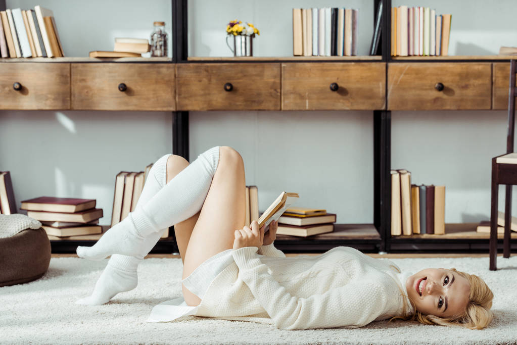 lächelnde schöne Frau, die auf Teppich liegt und Buch liest - Foto, Bild