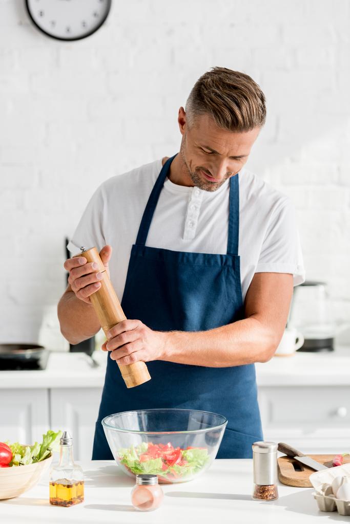 handsome adult man in apron salting salad at kitchen - Photo, Image