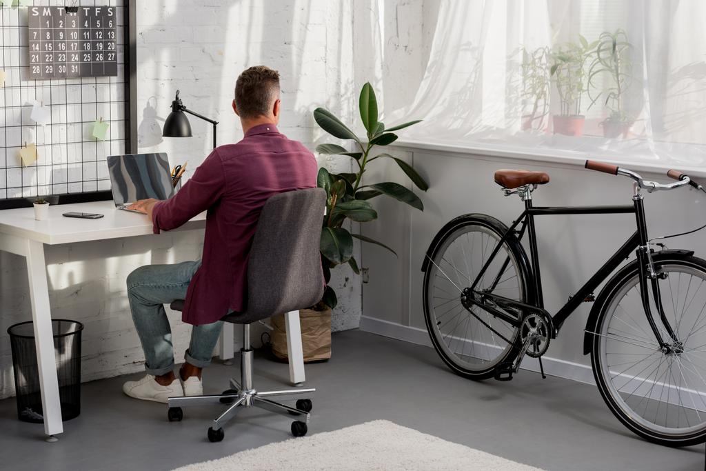vista trasera del hombre sentado en el lugar de trabajo y mirando por la ventana en la oficina en casa
 - Foto, Imagen