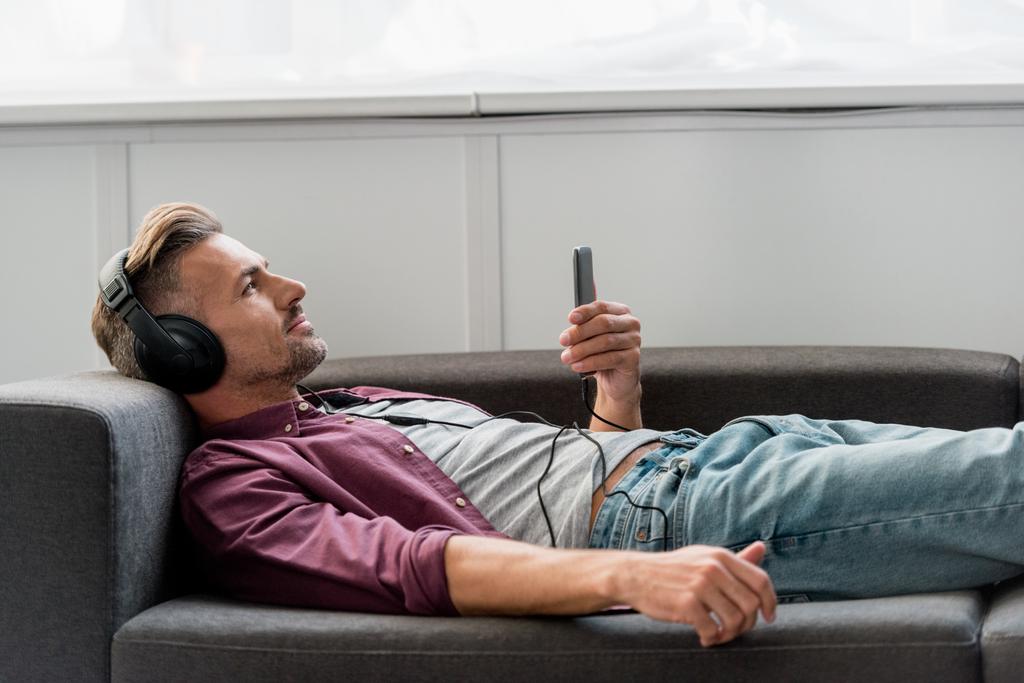 ευτυχισμένος άνθρωπος με smartphone ακούγοντας μουσική βάζοντας στον καναπέ  - Φωτογραφία, εικόνα