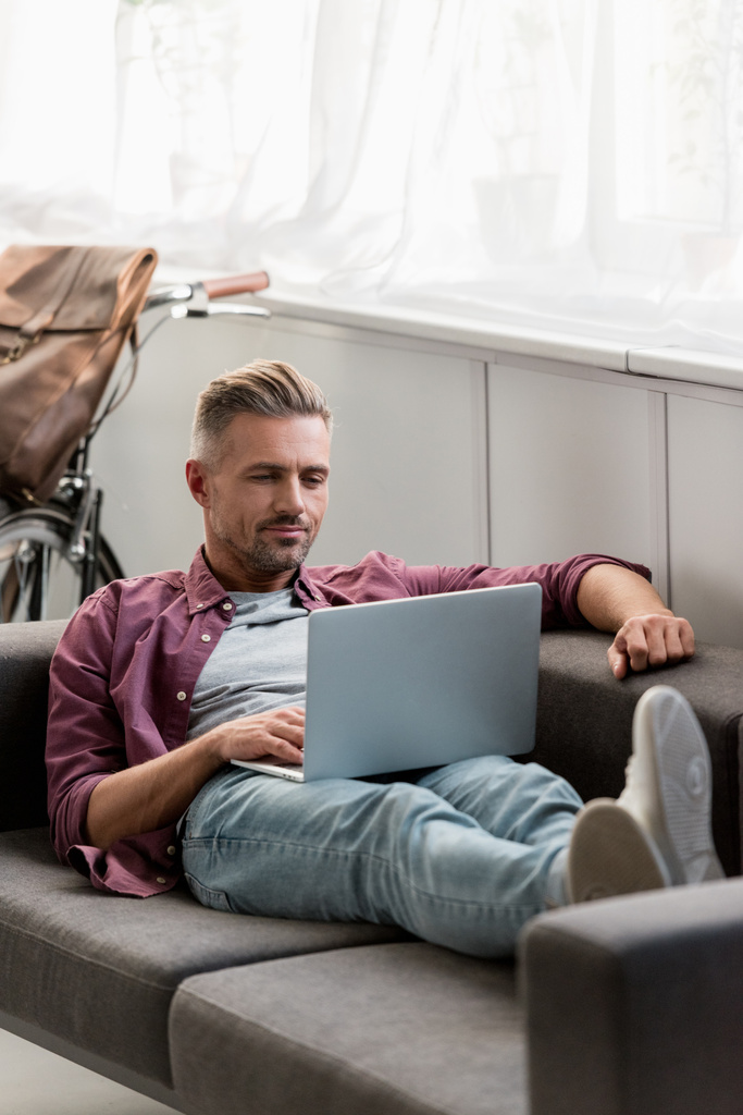 guapo freelancer acostado en el sofá y trabajando en el ordenador portátil en casa oficina
 - Foto, Imagen