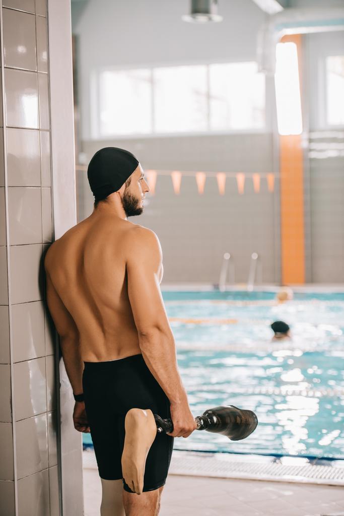 vista trasera del nadador parado junto a la piscina de la piscina cubierta y sosteniendo su pierna artificial
 - Foto, Imagen