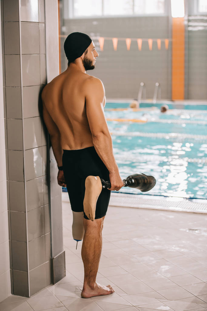 Rückansicht eines jungen Schwimmers, der am Beckenrand des Hallenbades steht und sein künstliches Bein hält - Foto, Bild