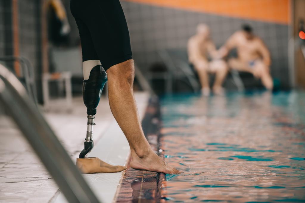 colpo ritagliato di sportivo con gamba artificiale in piedi a bordo piscina in piscina coperta e controllo della temperatura dell'acqua
 - Foto, immagini