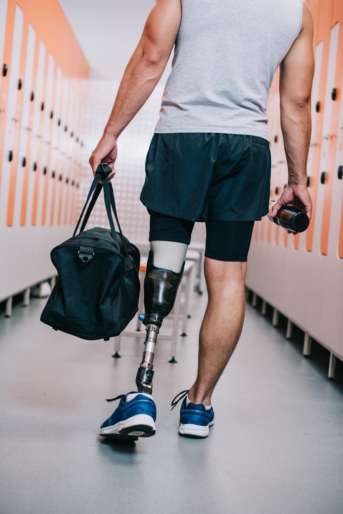 tiro recortado de deportista con pierna artificial de pie en el vestuario del gimnasio con botella y bolsa
 - Foto, Imagen