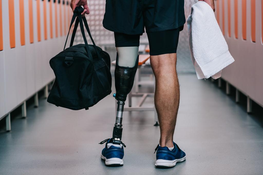 colpo ritagliato di sportivo con gamba artificiale in piedi in palestra spogliatoio con asciugamano e borsa
 - Foto, immagini