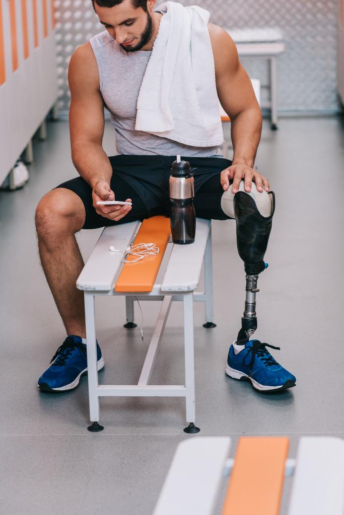 привлекательный молодой спортсмен с искусственной ногой сидит на скамейке в раздевалке спортзала и пользуется смартфоном
 - Фото, изображение