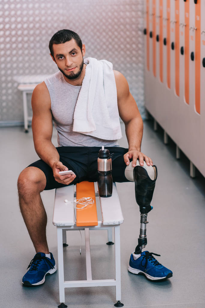 joven deportista guapo con pierna artificial sentado en el banco en el vestuario del gimnasio y el uso de teléfono inteligente
 - Foto, imagen