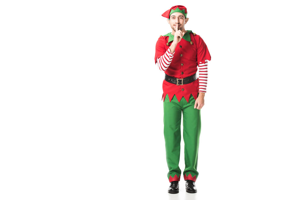 smiling man in christmas elf costume showing hush silence sign and looking at camera isolated on white - Photo, Image