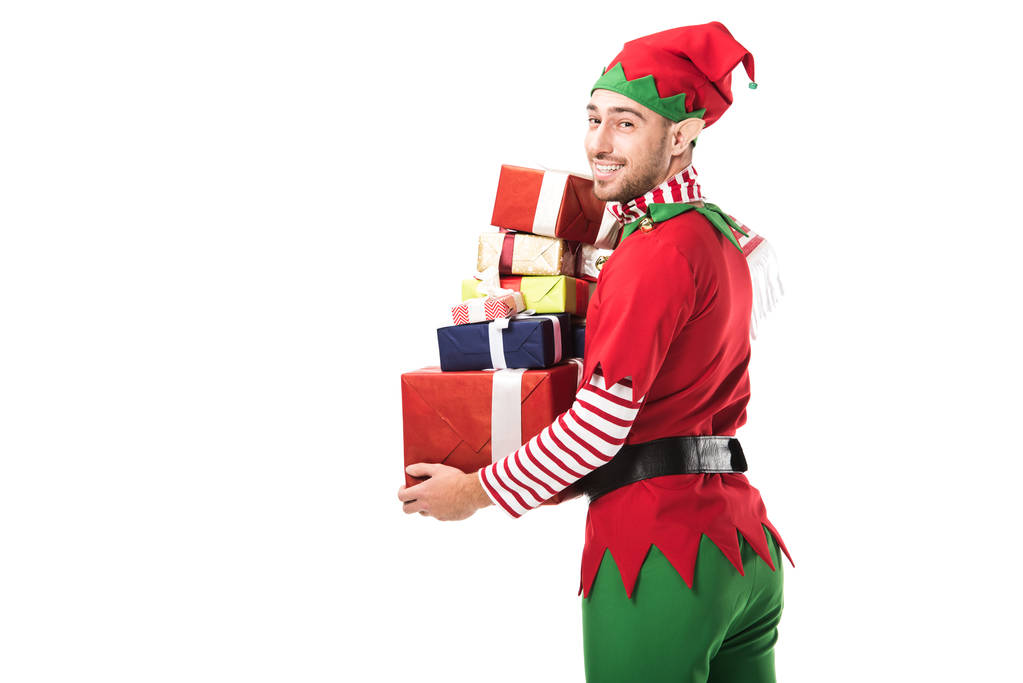 カメラ目線と白で隔離プレゼントの山を運ぶクリスマス エルフ衣装で笑みを浮かべて男 - 写真・画像