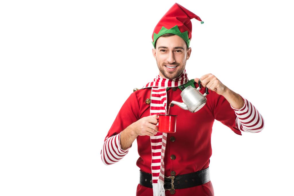 happy man in christmas elf costume with metal kettle pouring tea in cup and looking at camera isolated on white - Photo, Image