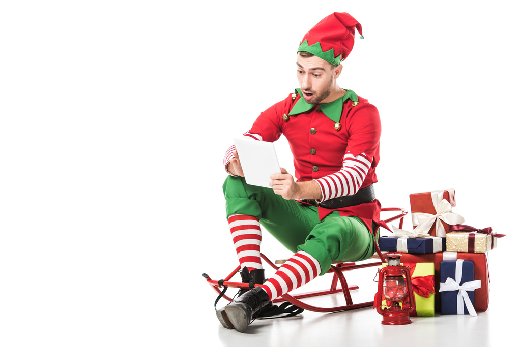 удивленный мужчина в рождественском костюме эльфа сидит на санях рядом с грудой подарков и смотрит на список желаний, изолированный на белом
 - Фото, изображение