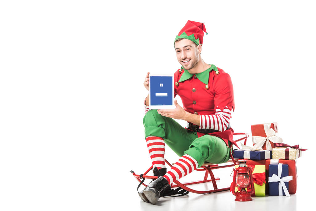 Mann im Weihnachtselfenkostüm sitzt auf Schlitten und hält Tablet mit Facebook-App auf dem Bildschirm - Foto, Bild