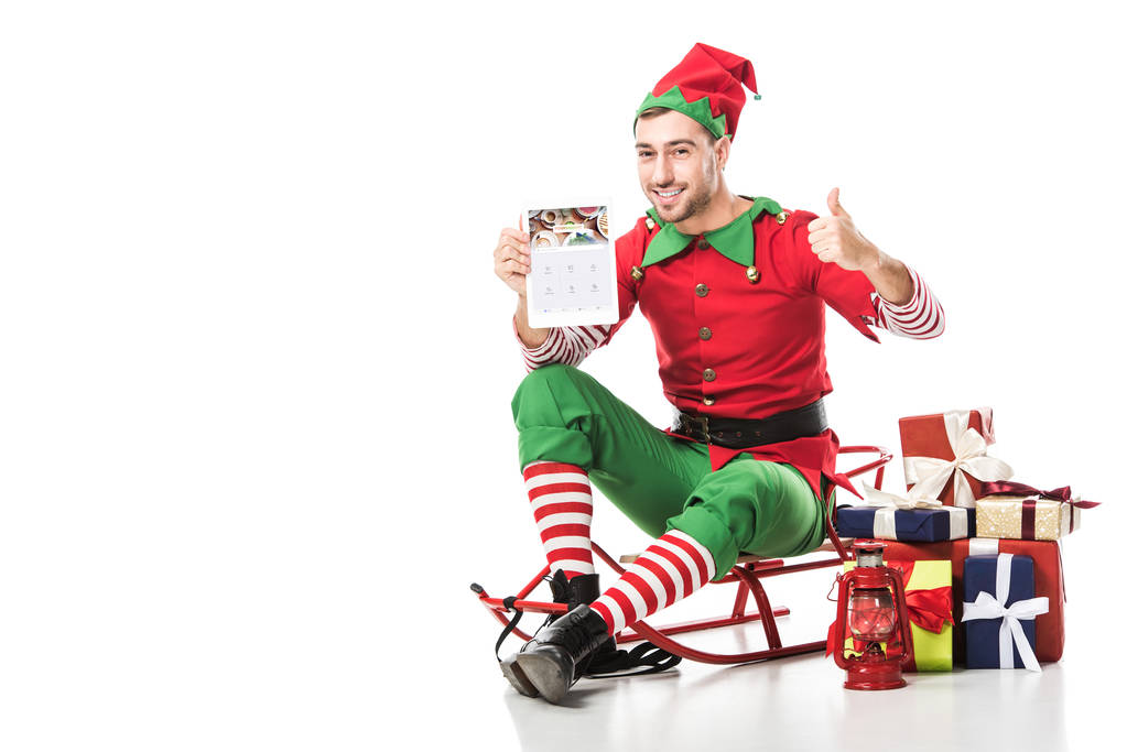 homem em traje de elfo de natal sentado no trenó, mostrando polegares para cima e segurando tablet com aplicativo forsquare isolado no branco
 - Foto, Imagem