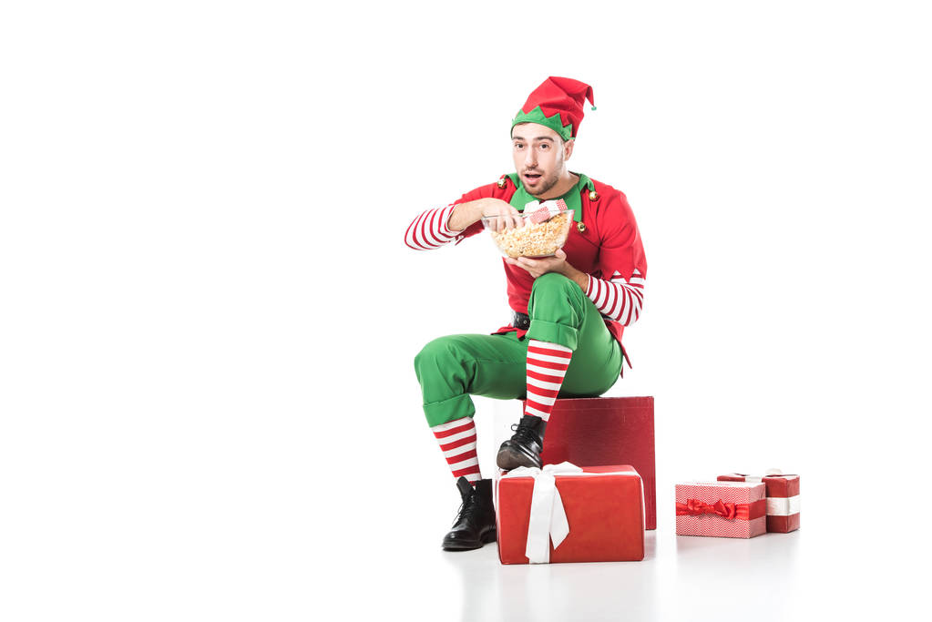 мужчина в рождественском костюме эльфа сидит на куче подарков и ест попкорн изолированный на белом
 - Фото, изображение