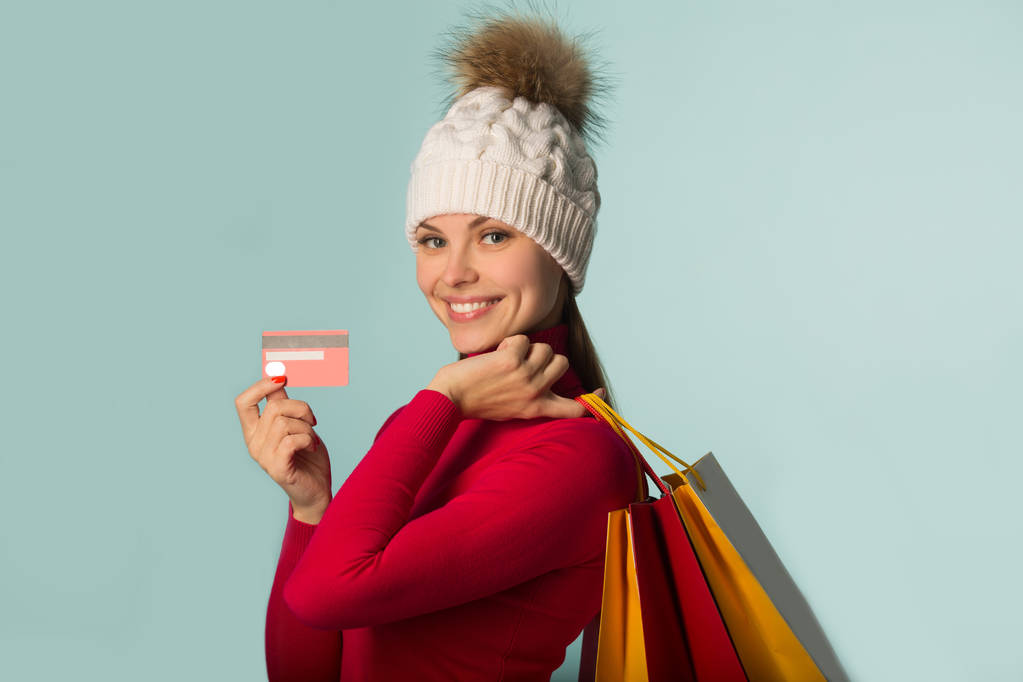 piękna młoda dziewczyna w kapeluszu zima trzyma karty płatniczej i pakiety w jej ręce na niebieskim tle - Zdjęcie, obraz