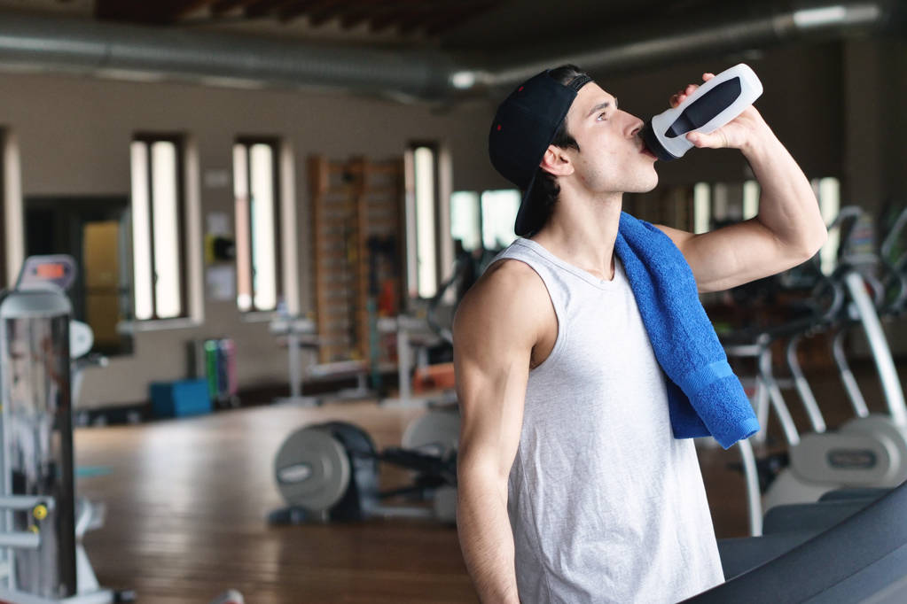 man during break drinking water from bottle while standing in gym  - Photo, Image