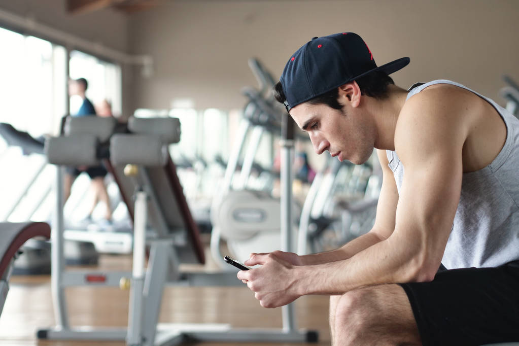 Portrait of a guy in a gym while resting between a workout and the other uses his phone to watch social networks or send messages. Concept of: sports, personal trainer, messages, telephone - Photo, Image