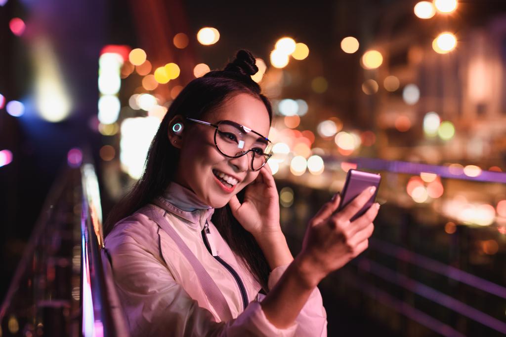 幸せなアジアの女の子のビデオ チャット、ネオンの光、未来の都市の通りのスマート フォンを使用して - 写真・画像
