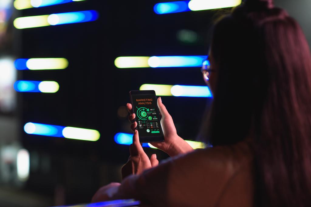 Widok z dziewczynka trzymając smartfon z marketingu urządzenie analizy na ulicy z neon światło wieczorem, miasto przyszłości koncepcja tyłu - Zdjęcie, obraz