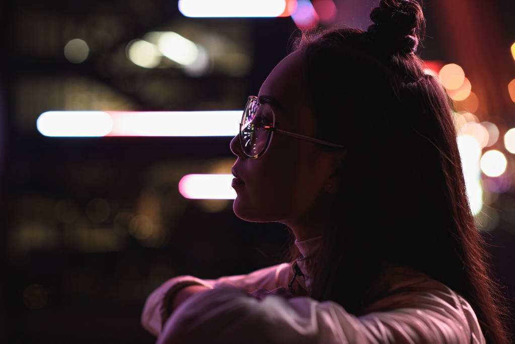 side view of stylish asian girl in glasses looking away on street with neon light in evening, city of future concept - Photo, Image