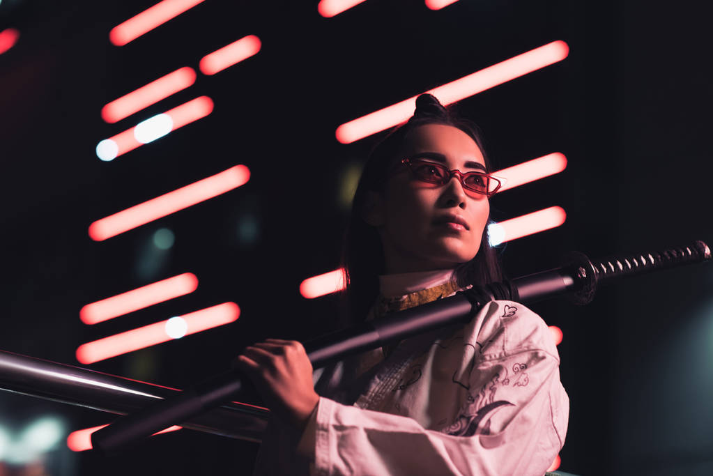 baixo ângulo vista de atraente asiático menina no branco quimono segurando japonês espada no rua com néon luz no noite, cidade de futuro conceito
 - Foto, Imagem