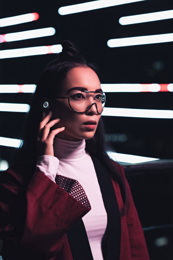 atractiva chica asiática en kimono de color burdeos y auriculares inalámbricos mirando hacia otro lado en la calle con luz de neón en la noche, ciudad del concepto futuro
 - Foto, Imagen