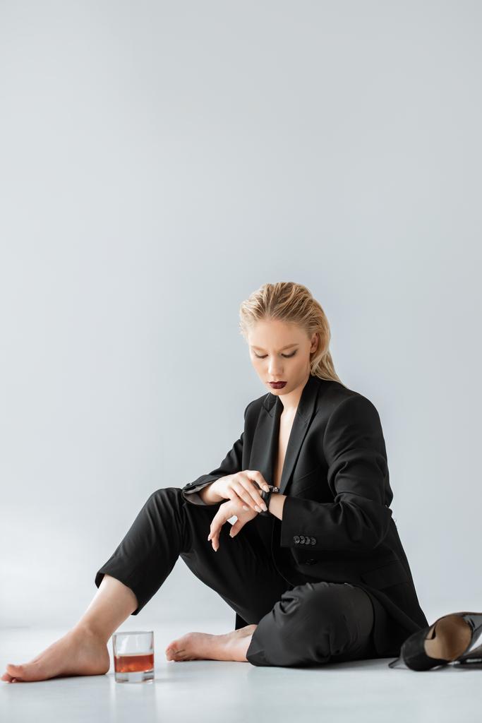 attraktives Mädchen im eleganten Anzug schaut auf die Uhr, während sie auf grau mit einem Glas Cognac und High Heels sitzt - Foto, Bild