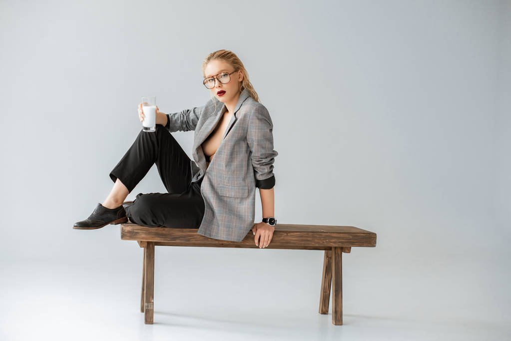 módní dívka v šedé sako držení sklenice mléka a sedí na dřevěnou lavici šedá - Fotografie, Obrázek