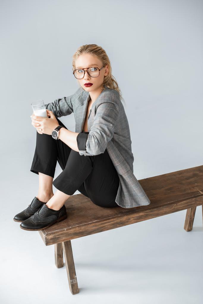 hermosa chica en chaqueta gris sosteniendo vaso de leche y sentado en banco de madera en gris
 - Foto, Imagen