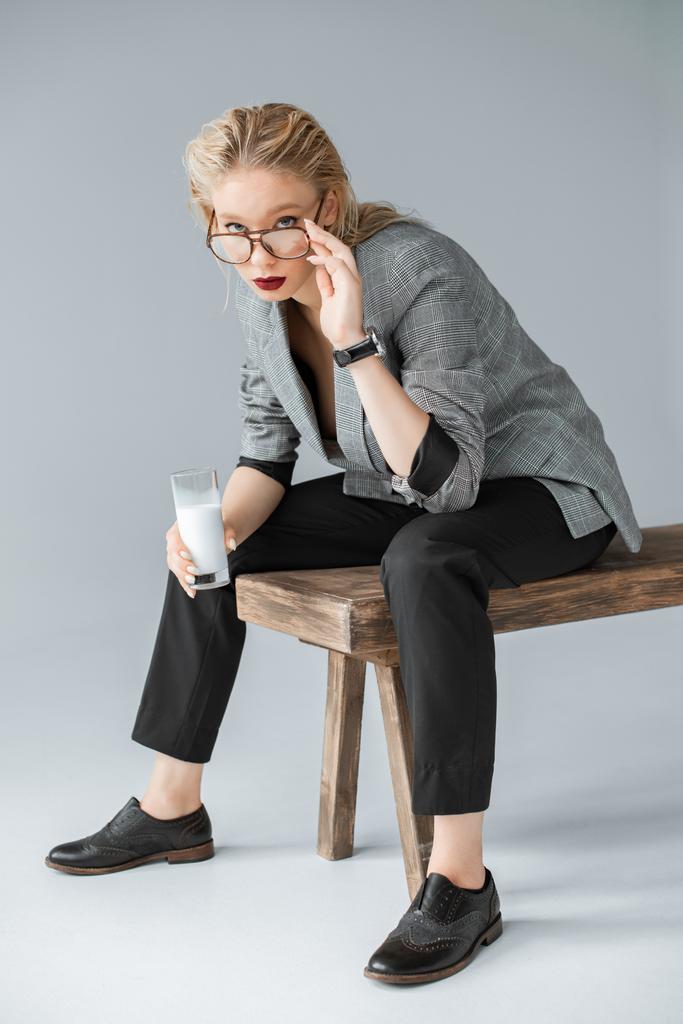 menina na moda em óculos segurando vidro de leite e sentado em banco de madeira em cinza
 - Foto, Imagem