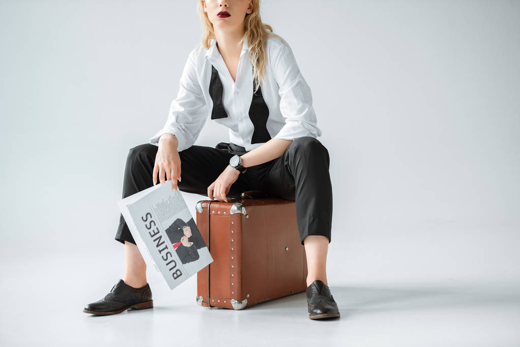 cropped view of stylish girl holding business newspaper while sitting on retro suitcase on grey - Photo, Image